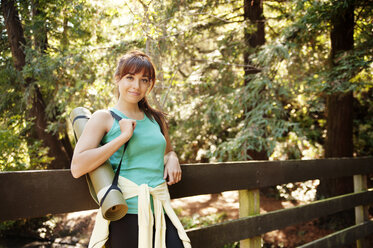 Portrait of confident woman carrying exercise mat while standing by railing in forest - CAVF06589