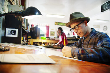 Mann schreibt auf Papier, während er am Schreibtisch in einer Werkstatt sitzt - CAVF06598