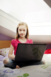Porträt eines Mädchens, das mit einem Laptop auf einem Bett zu Hause sitzt - CAVF06662
