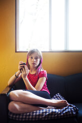 Porträt eines Mädchens, das ein Mobiltelefon benutzt, während es zu Hause auf dem Sofa sitzt - CAVF06812