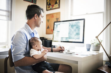 Mann trägt einen kleinen Jungen bei sich, während er zu Hause am Computer arbeitet - CAVF06882