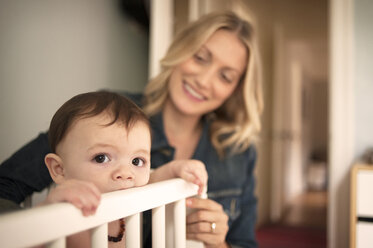 Mother looking at baby boy in crib at home - CAVF06906