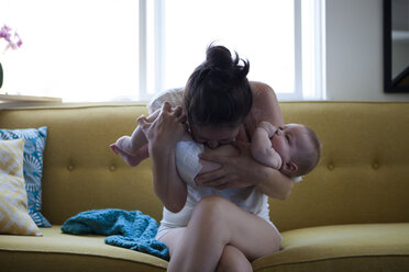 Mutter hält und küsst den Bauch des Babys, sitzt auf dem Sofa am Fenster - CAIF15326