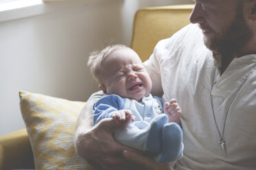 Father holding and looking at crying baby, sitting on sofa - CAIF15327