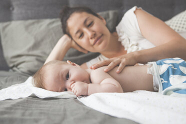 Porträt eines kleinen Babys, das am Daumen lutscht, mit lächelnder Mutter im Hintergrund - CAIF15341