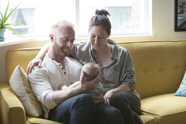 Eltern lächelnd und halten kleines Baby, sitzen auf gelbem Sofa - CAIF15343