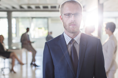 Porträt eines seriösen Geschäftsmannes mit Brille und Anzug, der in einem Büro steht, Kollegen im Hintergrund - CAIF15358
