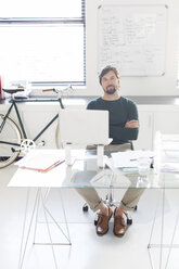 Portrait of man sitting with arms crossed behind glass desk in modern office, bicycle and whiteboard in background - CAIF15364