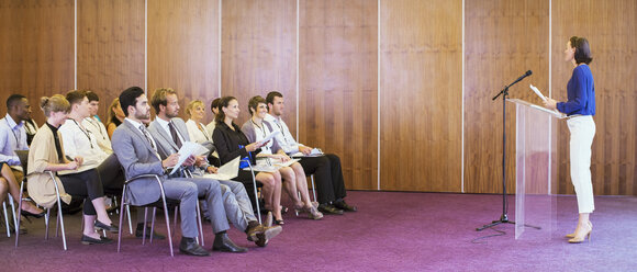 Young woman standing at transparent lectern, talking before audience in conference room - CAIF15476