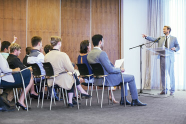 Portrait of businessman standing at transparent lectern talking before audience in conference room - CAIF15485