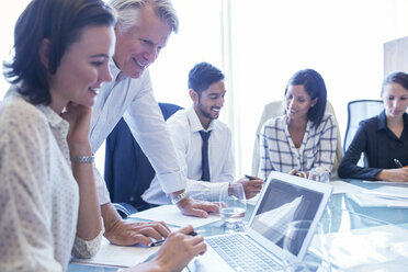 Geschäftsfrauen und Geschäftsmänner sitzen am Konferenztisch, benutzen einen Laptop und lächeln - CAIF15491