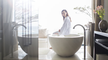 Portrait of smiling woman wearing white bathrobe, sitting on edge of bathtub in bathroom - CAIF15539