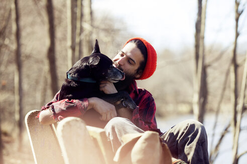 Affectionate man looking at dog while sitting on bench in forest during vacation - CAVF07017