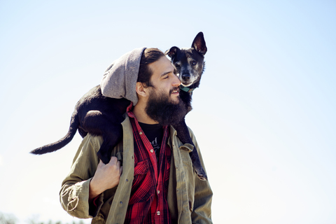 Nachdenklicher Mann, der einen Hund auf den Schultern trägt, während er vor einem klaren Himmel steht, lizenzfreies Stockfoto