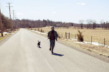 Mann und Hund laufen in voller Länge auf der Landstraße im Urlaub - CAVF07025