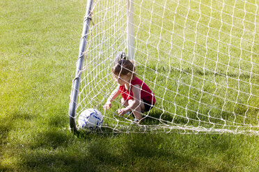 High angle view of girl playing with soccer ball while crouching in goal post on grassy field - CAVF07034