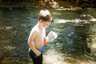 Side view of boy looking at fishing net while standing in river - CAVF07056