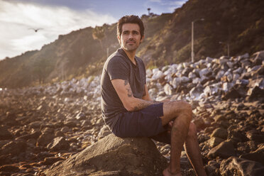 Thoughtful man looking away while sitting on rock at beach - CAVF07130