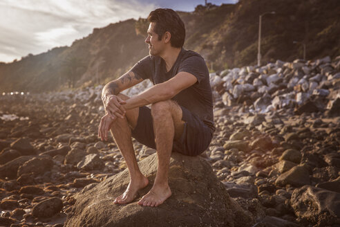 Mann schaut weg, während er auf einem Felsen am Strand sitzt - CAVF07131