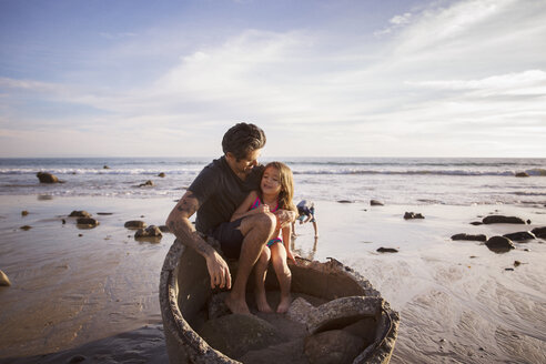 Father and daughter sitting on concrete pipe at beach during sunset - CAVF07136