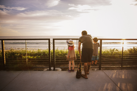 Rückansicht einer Familie, die während des Sonnenuntergangs an der Reling steht und auf das Meer blickt, lizenzfreies Stockfoto