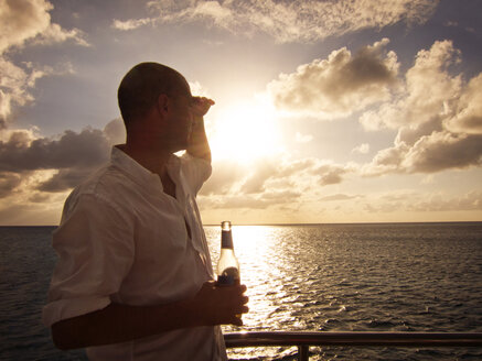 Man holding beer bottle while standing in yacht during sunset - CAVF07379