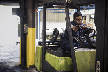 Portrait of manual worker showing peace signs while sitting in forklift at metal industry - CAVF07413