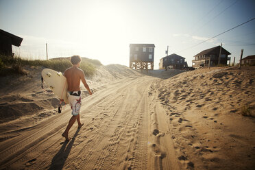 Rear view of man holding surfboard walking on sand at beach - CAVF07422