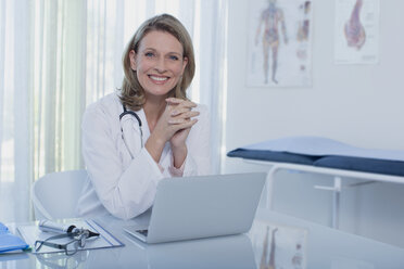 Porträt einer lächelnden Ärztin, die mit einem Laptop am Schreibtisch sitzt - CAIF15626