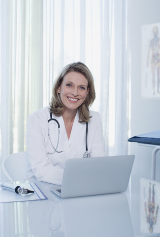 Porträt einer lächelnden Ärztin, die mit einem Laptop am Schreibtisch sitzt, lizenzfreies Stockfoto
