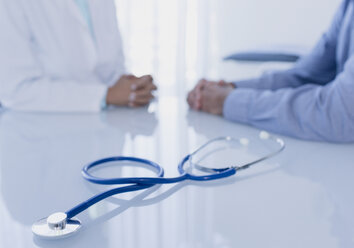 Stethoskop auf weißem Schreibtisch in einer Arztpraxis, Ärztin und Patientin sitzen im Hintergrund - CAIF15729