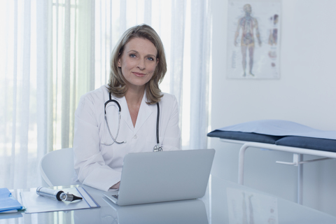 Porträt einer lächelnden Ärztin, die an einem Schreibtisch mit Laptop im Büro sitzt, lizenzfreies Stockfoto