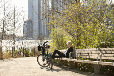 Woman resting on bench besides parked bicycle by river in city - CAVF07521