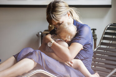 Mutter, die ihre Tochter umarmt, während sie auf einem Liegestuhl zu Hause sitzt - CAVF07559