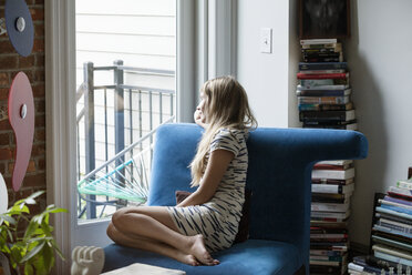 Frau schaut durch das Fenster, während sie zu Hause auf dem Sofa sitzt - CAVF07572