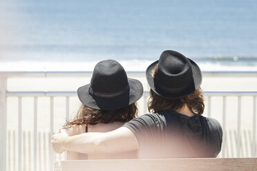 Rückansicht eines Paares mit Hüten, das auf einer Bank am Strand sitzt - CAVF07594