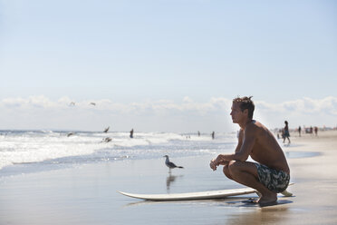 Nachdenklicher Mann mit Surfbrett am Strand hockend - CAVF07616