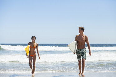Glückliches Paar, das ein Surfbrett trägt, während es am Strand gegen den klaren Himmel läuft - CAVF07644