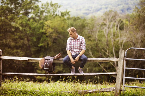 Mann schaut auf den Sattel, während er auf dem Geländer eines Bauernhofs sitzt - CAVF07653