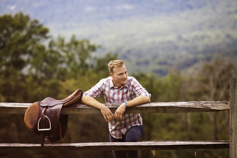 Glücklicher Rancher mit Sattel am Zaun lehnend auf einem Bauernhof, lizenzfreies Stockfoto