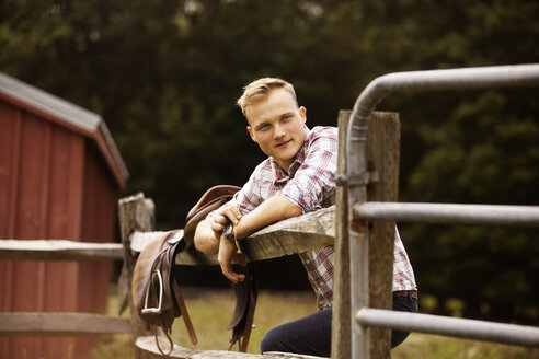 Glücklicher Rancher mit Sattel am Zaun lehnend auf einem Bauernhof - CAVF07656