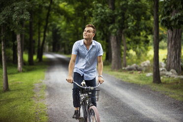 Happy man looking up while riding bicycle on road in woodland - CAVF07677
