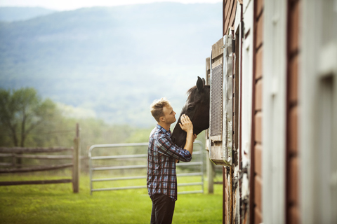 Rancher streichelt Pferd und schaut durch den Stall, lizenzfreies Stockfoto