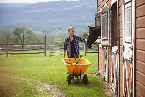 Rancher streichelt Pferd beim Schieben einer Schubkarre auf einem Bauernhof - CAVF07683