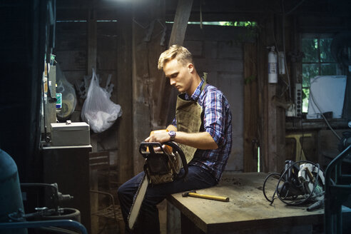 Mann hält Kettensäge, während er auf einem Tisch in einer Werkstatt sitzt - CAVF07686