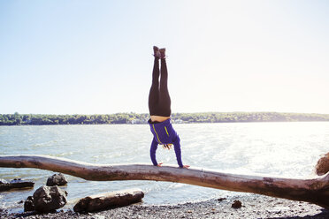 Woman doing handstand on log at riverbank against clear sky - CAVF07738