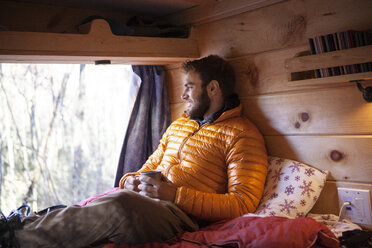 Lächelnder Mann mit Becher, der wegschaut, während er auf dem Bett im Wohnmobil sitzt - CAVF07791