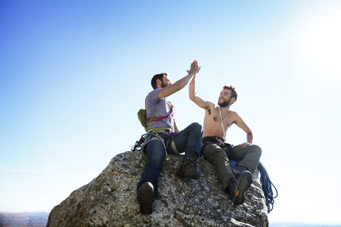 Glückliche Freunde, die sich auf einem Felsen sitzend gegen den klaren Himmel ein High-Five geben - CAVF07809