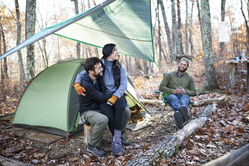 Freund schaut Paar an, während er auf Baumstümpfen bei einem Zelt im Wald sitzt - CAVF07831