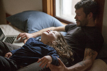 Boy looking at father using laptop computer while relaxing on bed at home - CAVF07883